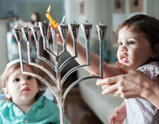 ילדה מדליקה נר חנוכה עם ליווי אחד ההורים